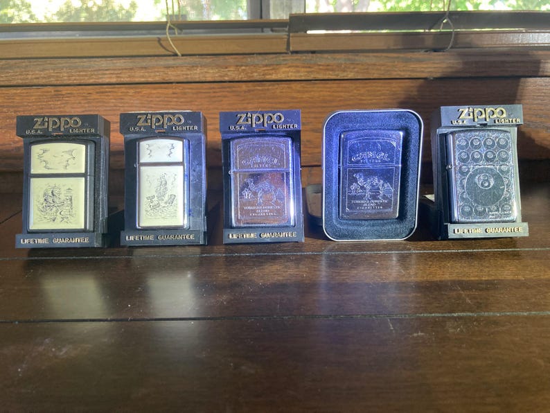 May include: Five Zippo metal lighters displayed in individual packaging. Four lighters are housed in black plastic stands labelled Zippo U.S.A. Lighter and Lifetime Guarantee, while one is in a silver-rimmed case. Each features unique engravings.