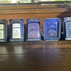 May include: Five Zippo metal lighters displayed in individual black plastic stands. Each lighter features a unique engraved design on the front surface, including Camel cigarette branding and a billiard ball motif, all marked with the Zippo logo.