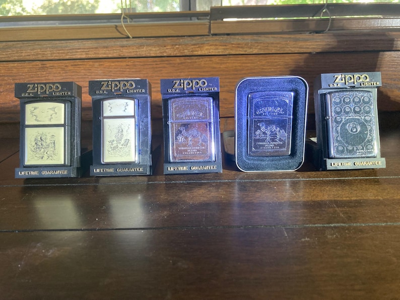 May include: Five Zippo metal lighters displayed in individual black plastic stands. Each lighter features a unique engraved design on the front surface, including Camel cigarette branding and a billiard ball motif, all marked with the Zippo logo.