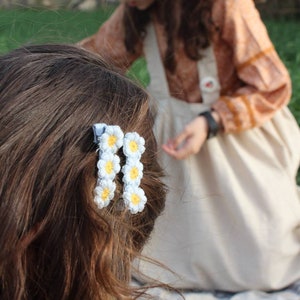 Può includere: Primo piano dei capelli castani adornati da due fermagli per capelli a forma di margherita bianca e gialla. Le clip sono attaccate a un nastro azzurro. Sullo sfondo, una persona indossa un vestito beige.
