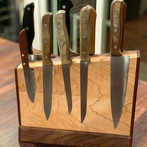 May include: A wooden knife block with a walnut base and a maple top. Seven knives are inserted into the block, including a paring knife, a chef's knife, and a bread knife.
