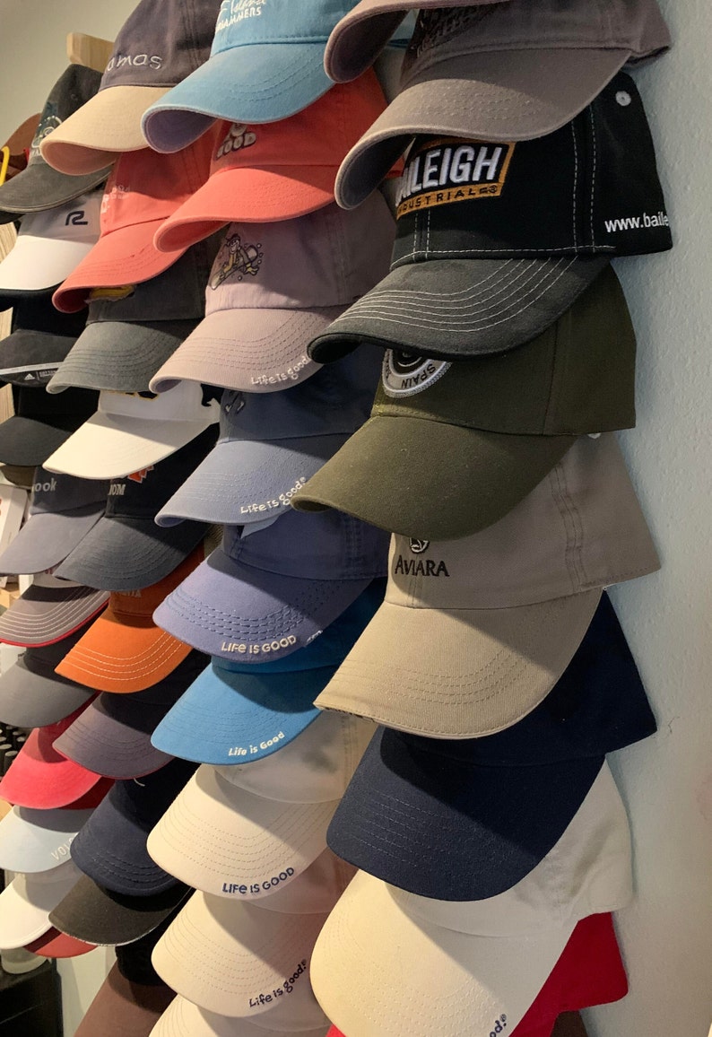 May include: A wall display of various baseball caps, including several with the text "Life is Good" embroidered on them. Some caps have logos, including "Raleigh Industrials" and "Aviara".