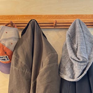May include: A horizontal wooden wall-mounted coat rack featuring alternating light and dark wood grain stripes with five upward-curving hooks. The rack holds an orange and gray baseball cap with an Acadia National Park patch, a dark olive jacket, and a gray beanie.