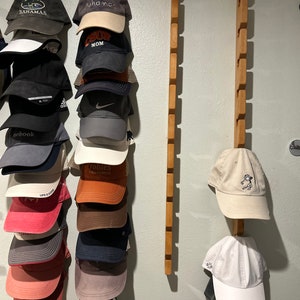 May include: A wall mounted hat rack with two wooden rails. The rails are filled with a variety of baseball caps in different colors and designs. The caps include brands like "Life is Good", "Trobbies California", "Bahamas", and "Surf Mom".
