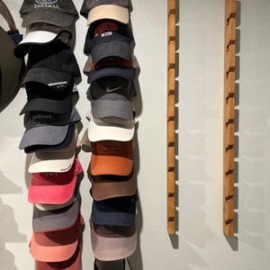 May include: A wall mounted hat rack with 14 slots, holding 13 baseball caps and 1 visor. The caps are in various colors and designs, including a cap with the word "Bahamas" on it, a cap with the word "Surf Mom" on it, and a cap with the word "Pebblebrook" on it.