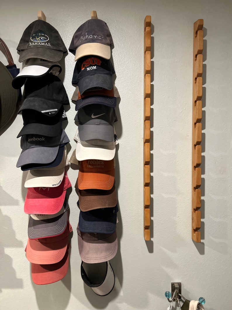 May include: A wall mounted hat rack with 14 slots, holding 13 baseball caps and 1 visor. The caps are in various colors and designs, including a cap with the word "Bahamas" on it, a cap with the word "Surf Mom" on it, and a cap with the word "Pebblebrook" on it.