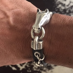 May include: A black leather bracelet with a silver horse head clasp. The clasp is detailed with a ring and a small chain.