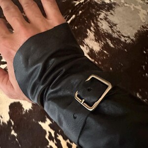 May include: A black leather wrist cuff featuring a gathered, ruched texture and a polished silver-tone metal buckle closure. The cuff wraps around the wrist, with the strap threaded through the buckle to secure the fit against the skin.