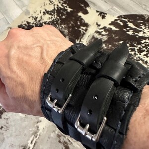 May include: A wide black leather cuff bracelet featuring a textured surface and two adjustable straps with silver-toned metal buckles. The cuff is constructed with multiple overlapping leather segments and heavy stitching along the edges.