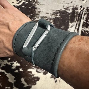 May include: A wide, dark gray leather cuff bracelet featuring a silver-toned metal carabiner attached to the front. The cuff has visible stitching along the edges and wraps securely around the wrist, showcasing a textured, matte finish on the leather.