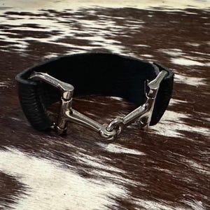 May include: A black leather cuff bracelet featuring a polished silver-tone metal horse bit connector. The wide band is constructed from smooth black leather and is secured by a metallic equestrian-inspired bit hardware piece at the centre.