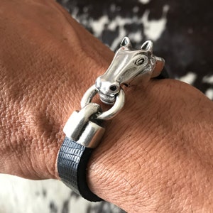 May include: A bracelet featuring a textured black leather strap and a polished silver-tone metal horse head charm. The horse head is connected to the strap by a silver-tone metal ring, creating a secure clasp mechanism for the wrist accessory.
