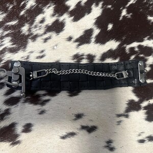 May include: A black leather cuff bracelet featuring a textured, grid-like pattern. The bracelet is secured with a silver-toned metal latch mechanism on one side and a thin, delicate silver-toned chain that connects across the center.