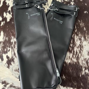 May include: A pair of black leather half chaps featuring a full-length silver metal zipper, adjustable buckle straps at the top, and a small silver chain detail. The text WICKED PONY by Chlo is printed in white on the upper portion of each chap.