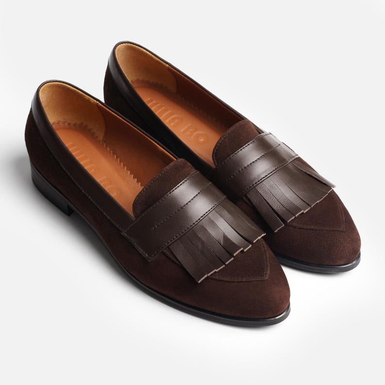 Pair of chocolate-brown suede women’s loafers with smooth leather penny straps, layered kiltie fringe, almond toes and low black heels.