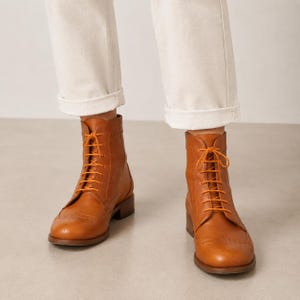 May include: A pair of tan leather ankle boots with orange laces. The boots have a classic brogue design with decorative perforations and a low heel. The laces are tied in a neat bow.