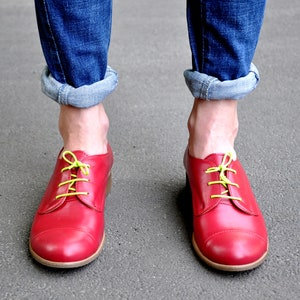 Könnte beinhalten: Rote Lederschuhe mit gelben Schnürsenkeln. Die Schuhe werden von einer Person getragen, die blaue Jeans trägt.