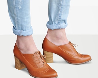 May include: Brown leather lace-up shoes with a block heel. The shoes have a wingtip design and are worn with rolled-up blue jeans.