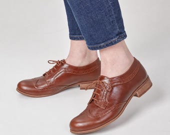 May include: A pair of brown leather oxford shoes featuring classic wingtip brogue detailing, a rounded toe, thin matching laces, and a low stacked heel, worn with dark blue denim jeans that end just above the ankle for a clean, polished look.