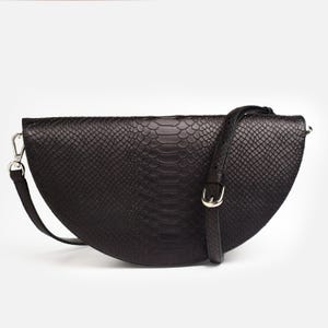 May include: A black, semi-circular crossbody bag featuring a textured snakeskin pattern. The bag includes an adjustable shoulder strap with a silver-toned metal buckle and a matching silver-toned metal clasp on the side for attachment.