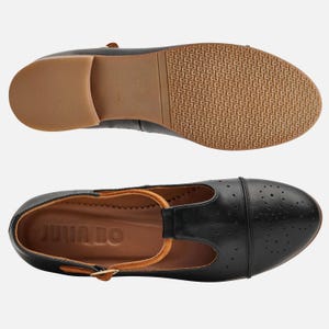 May include: Black leather t-strap shoes with a perforated design and a brown leather insole. The shoes have a brown leather strap with a buckle and the words "Julia Bo" are printed on the insole.