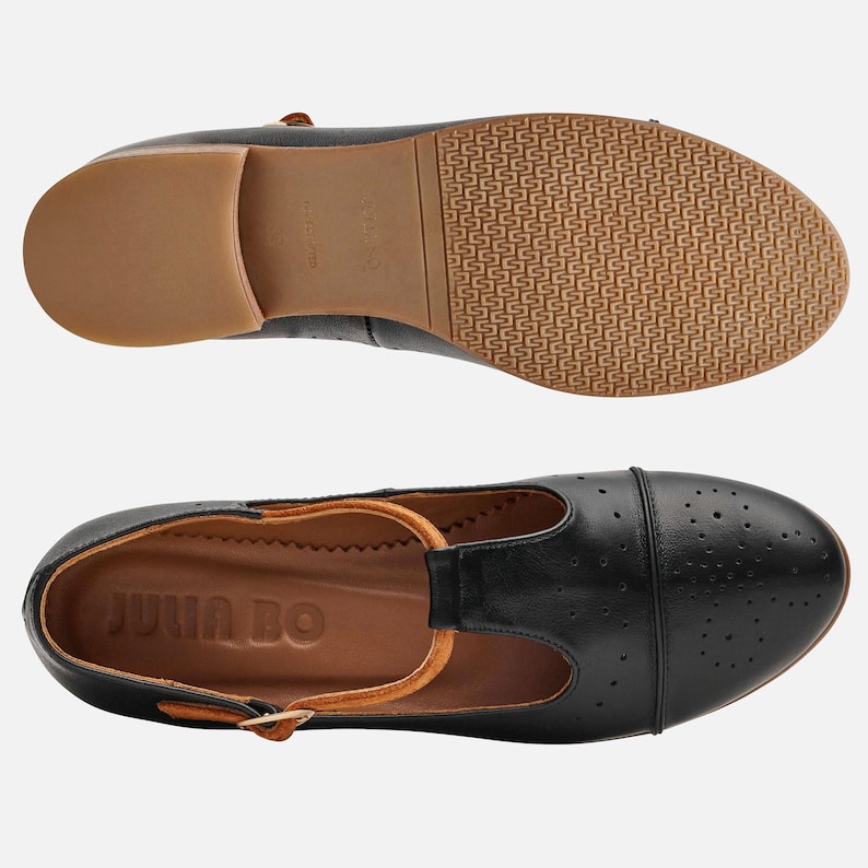 May include: Black leather t-strap shoes with a perforated design and a brown leather insole. The shoes have a brown leather strap with a buckle and the words "Julia Bo" are printed on the insole.