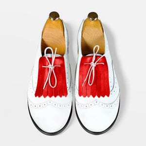 May include: A pair of white leather shoes with red fringe detailing. The shoes have a classic Oxford design with decorative perforations along the edges and black soles. The shoes are displayed on wooden shoe trees.