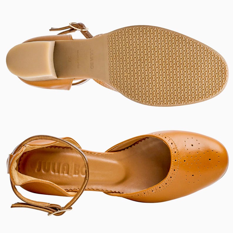 Puede incluir: Dos pares de zapatos de cuero color marr&oacute;n claro con correas en el tobillo y tacones peque&ntilde;os. Los zapatos tienen un dise&ntilde;o cl&aacute;sico con perforaciones decorativas en la puntera. Las suelas son de color marr&oacute;n claro con un patr&oacute;n texturizado. La marca "JULIA BO" es visible.