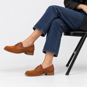 May include: A pair of brown suede loafers featuring smooth leather trim around the collar, a moccasin-style stitched toe, thin brown laces, gold-tone eyelets, and a low stacked heel. The shoes are worn with dark blue denim jeans and a black blazer.