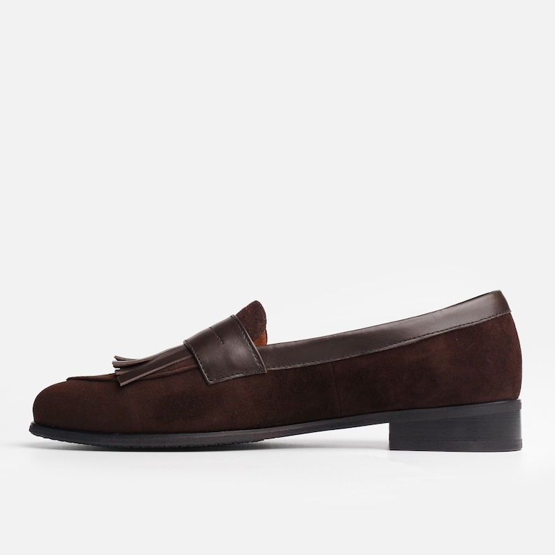 May include: A dark brown suede loafer featuring a smooth leather saddle strap across the vamp, a decorative tassel detail, a low block heel, and a dark brown leather trim along the collar. The shoe has a rounded toe and a flat black outsole.