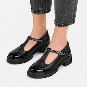 May include: Black leather T-strap shoes with a glossy finish on the toe and a chunky sole. The strap features a dotted pattern and a small buckle. The shoes have a classic brogue design.