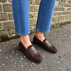 Woman wearing chocolate-brown suede kiltie loafers with straight cropped blue jeans beside a brick wall.