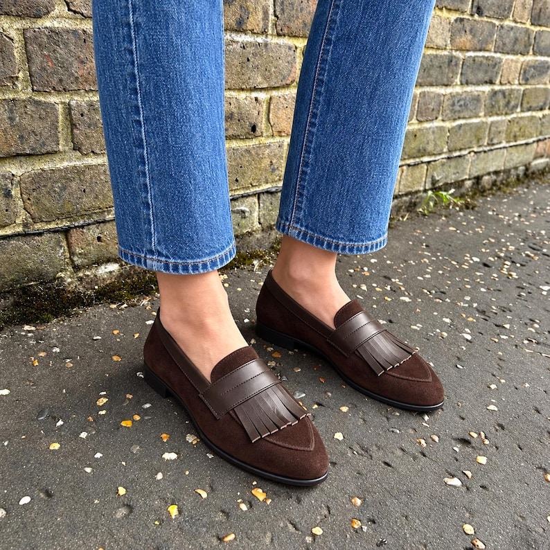 Woman wearing chocolate-brown suede kiltie loafers with straight cropped blue jeans beside a brick wall.
