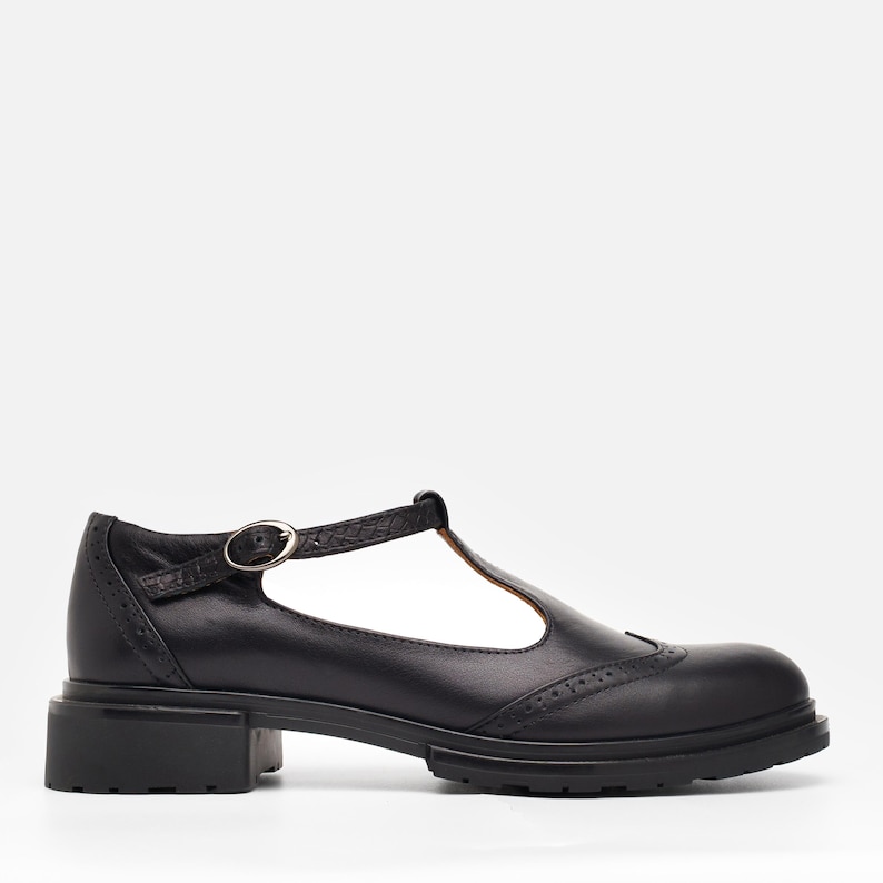 May include: A black leather T-strap shoe featuring a rounded toe, decorative brogue perforations along the toe cap and heel, a thin adjustable ankle strap with a silver-toned metal buckle, and a thick, lugged black rubber sole with a low block heel.
