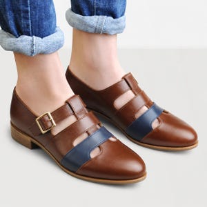 May include: A pair of brown leather shoes with a cutout design, featuring a navy blue strap across the vamp and a gold-toned metal buckle closure on the side. The shoes have a low heel and a rounded toe, providing a classic look for footwear.