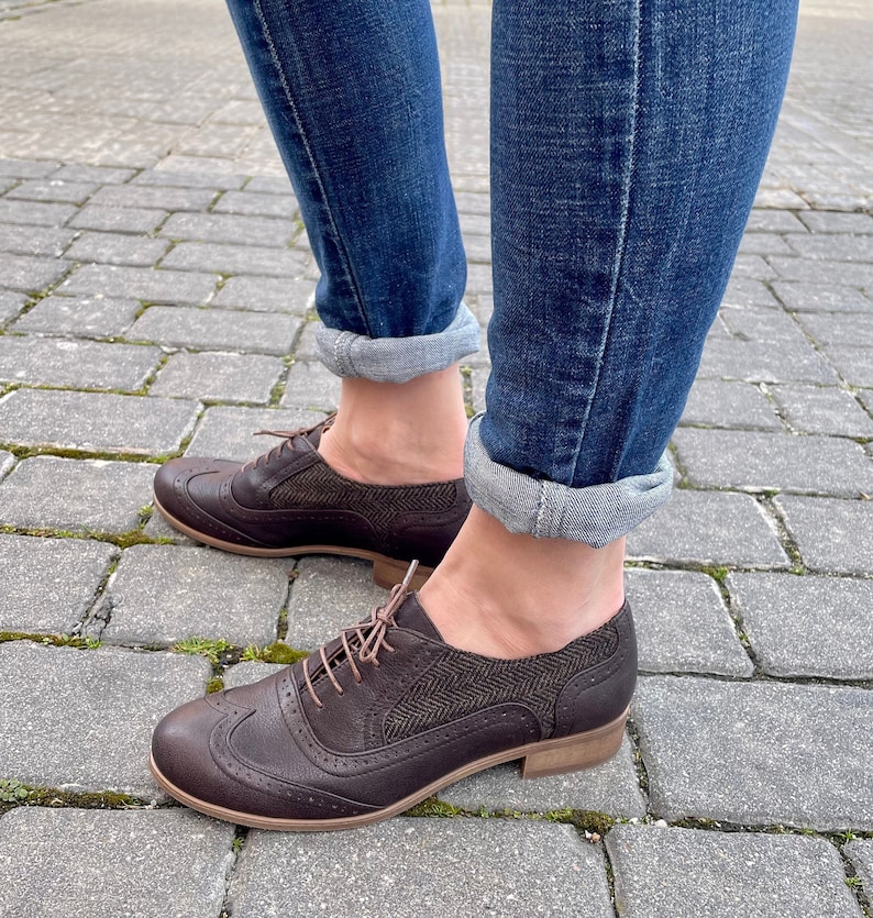May include: Brown leather oxford shoes with a tweed detail on the side. The shoes are worn with rolled up blue jeans.