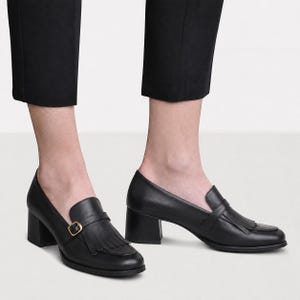 May include: A pair of black leather loafers with a block heel, featuring a fringe detail on the vamp and a thin strap across the top secured with a small gold-toned metal buckle. The shoes have a rounded toe and a smooth, matte finish.