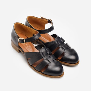 May include: A pair of black leather sandals with a closed toe design and ankle straps. The sandals have a low heel and a gold-coloured buckle. The interior of the sandals is brown.