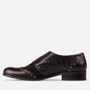 May include: Black leather shoe with a low heel. The shoe features a textured, crocodile-like pattern on the strap and a suede panel. Small silver studs adorn the toe and heel areas, adding a touch of detail to the design.