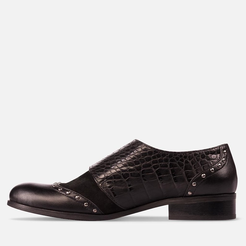 May include: Black leather shoe with a low heel. The shoe features a textured, crocodile-like pattern on the strap and a suede panel. Small silver studs adorn the toe and heel areas, adding a touch of detail to the design.