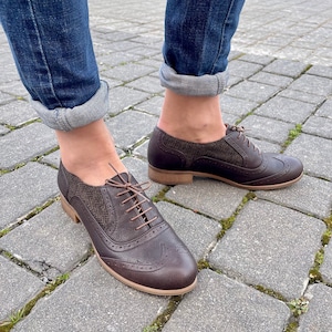 May include: Brown leather oxford shoes with a tweed detail on the back. The shoes have a lace-up closure and a wingtip design.