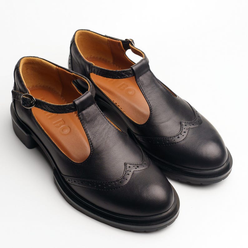 May include: A pair of black leather T-strap shoes featuring a rounded toe, decorative brogue perforations along the wingtip seam, a thin adjustable ankle strap with a metal buckle, and a tan interior lining with the embossed text BAMBO.