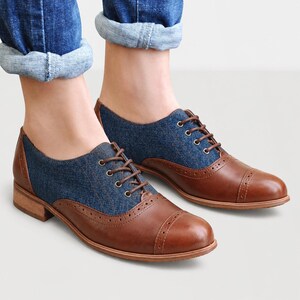 May include: Brown and blue oxford shoes with a cap toe and a denim-like fabric on the upper. The shoes have brown laces and a low heel. The shoes are worn with blue jeans.