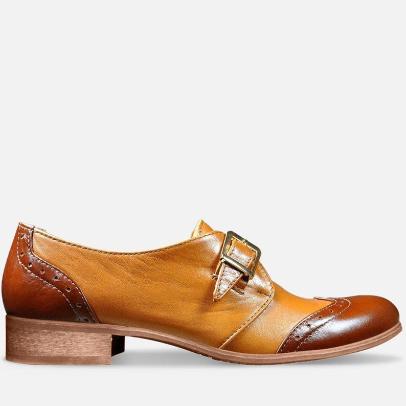 May include: Tan leather women's shoes with a brown leather toe cap and a single buckle strap. The shoes have a low block heel.