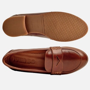 May include: A pair of brown leather loafers with a buckle detail. The loafers have a brown leather sole and the brand name "JULIA BO" is embossed on the insole.
