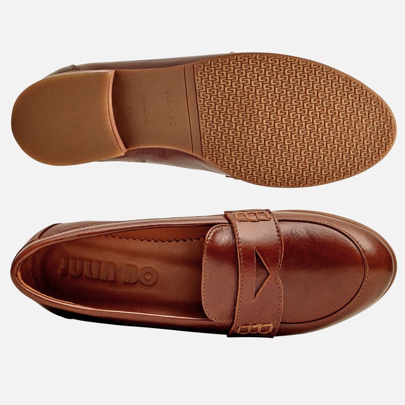 May include: A pair of brown leather loafers with a buckle detail. The loafers have a brown leather sole and the brand name "JULIA BO" is embossed on the insole.