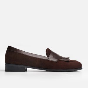 May include: A dark brown suede loafer featuring a smooth leather saddle strap across the vamp, a decorative tassel detail, a rounded toe, and a low black block heel. The shoe is constructed with a combination of textured suede and smooth leather.