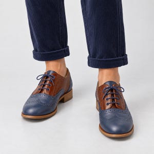 May include: A pair of two-tone oxford shoes featuring a brown leather upper and a blue leather toe cap with decorative brogue detailing. The shoes have blue laces, a low stacked heel, and are worn with dark blue cuffed corduroy trousers.