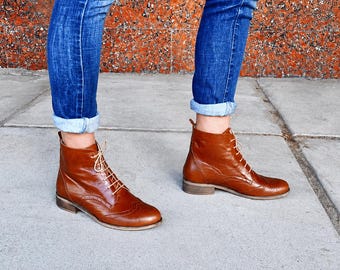 May include: A pair of brown leather ankle boots with light-colored laces. The boots have a low heel and a classic design. The person is wearing blue jeans with the cuffs rolled up, showcasing the boots.
