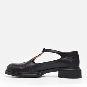 May include: A black leather T-strap shoe featuring a rounded toe, decorative brogue perforations along the vamp, and a textured strap. The shoe is constructed with a low, block heel and a thick, lugged rubber sole for a sturdy, flat profile.